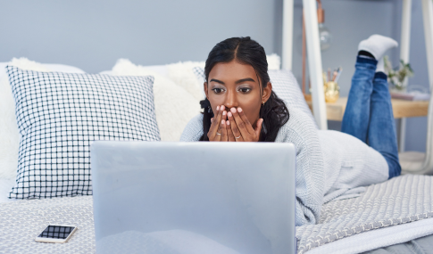 Image of a girl sitting on a bed with her hands over her mouth in shock while looking at her laptop