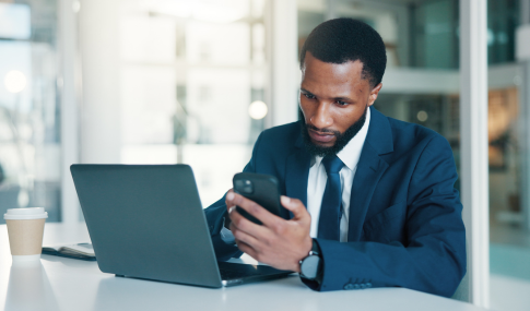 Man sitting at a desk in front of a laptop while holding a cellphone