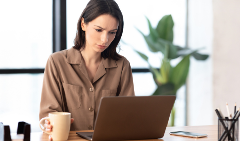 Woman holding her coffee mug, looking at her laptop with a questioning look on her face