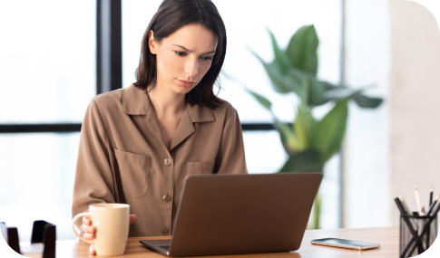 Image of a woman working on her laptop, with her hand on a coffee cup, while sitting at a desk