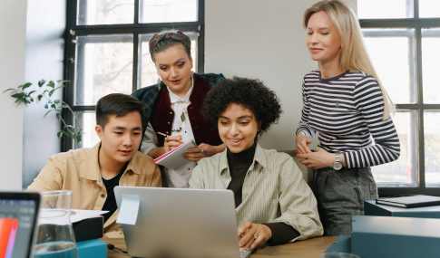 Image of 4 people looking at a laptop, one woman writing down information in a notepad