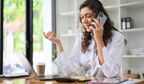 Image of lady on the cell phone while looking at her laptop and sitting at a desk