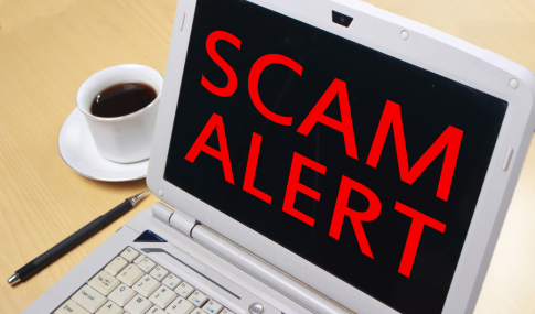 Image of a laptop with the words Scam Alert sitting on a desk next to a cup of coffee