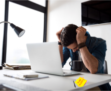 Man with head in hands at a desk in front of laptop with a sticky note that says Scam Alert