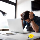 Man with head in hands at a desk in front of laptop with a sticky note that says Scam Alert