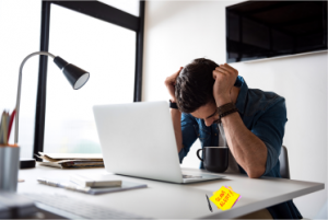 Man with head in hands at a desk in front of laptop with a sticky note that says Scam Alert